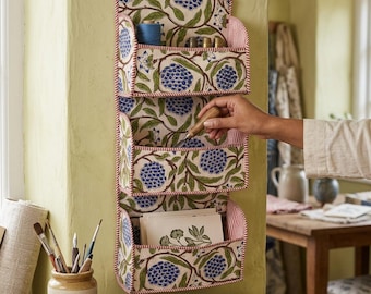 Op de afbeelding: Een stoffen wandorganizer met drie vakken, voorzien van een crèmekleurige achtergrond met een herhalend patroon van blauwe hortensia-achtige bloemen en groene bladeren. De randen zijn afgewerkt met rood-wit gestreepte stof. Bevat naai-artikelen.