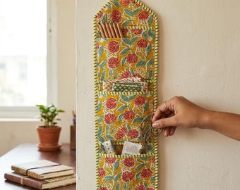 Op de afbeelding: Een verticale stoffen wandorganizer met een gele achtergrond en een herhalend bloemenpatroon in rood en blauwgroen. De organizer heeft drie gelaagde vakken met potloden, kleine stukjes stof, een schaar en witte papieren kaarten.