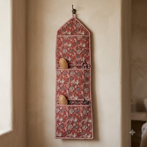 May include: A quilted wall hanging organiser featuring a rust-coloured fabric with a dark floral pattern and red and white striped trim. The vertical organiser has three open pockets, each containing a wooden hairbrush and metal scissors.