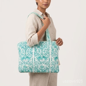 Puede incluir: Un bolso tote grande con un estampado floral turquesa y blanco. El bolso tiene un cierre de cremallera y asas a rayas turquesas y blancas a juego. El bolso está hecho de tela acolchada.
