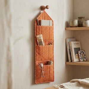 May include: A vertical fabric wall organiser featuring an orange and white gingham pattern with three tiered pockets. The organiser hangs from a wooden peg and contains a notebook, a small package of incense sticks, and a pair of reading glasses.
