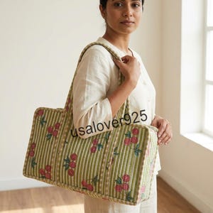 May include: A rectangular tote bag featuring a light green and white vertical striped pattern with a repeating print of small red cherries and green leaves. The bag has two shoulder straps and a structured shape with visible quilted stitching.
