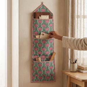 May include: A vertical fabric wall organiser featuring three tiered pockets with a repeating pattern of pink flowers and green stems on a light green background, finished with red and white striped trim and a pointed top hanging loop.