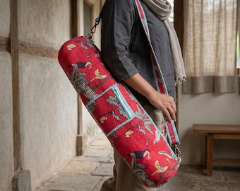 May include: A cylindrical red yoga mat bag featuring a tropical print with toucans and palm leaves. The bag has a long, adjustable shoulder strap with a matching red and teal striped pattern, secured by metal clips on each end of the carrier.