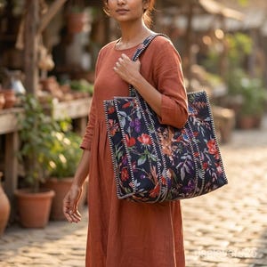 May include: A woman carries a large tote bag with a floral pattern. The bag is black with a colorful design of red, purple, and green flowers and leaves. The bag has a white and black striped border and matching straps.