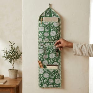 Può includere: Un organizer da parete verticale in tessuto con motivo floreale verde e bianco. Presenta tre tasche sovrapposte contenenti un taccuino, occhiali, chiavi e matite. I bordi sono rifiniti con un profilo a righe verdi e bianche.