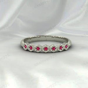 Peut inclure: Une bague en argent ornée d'une série de sertissages hexagonaux, chacun contenant une pierre précieuse rouge vif. L'anneau de la bague est lisse et poli, et les pierres précieuses sont espacées uniformément. La bague est présentée sur une surface blanche texturée.