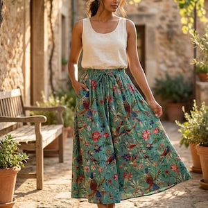 May include: A woman wearing a cream-colored tank top and a teal skirt with a floral and bird print. The skirt has a drawstring waist and pockets. The background features a stone building and greenery.