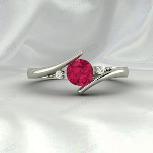 Peut inclure: Une bague en argent avec une grosse pierre rouge ronde au centre. La bague comporte deux petites pierres blanches de chaque côté de la pierre centrale. Le groupe a un design unique et fluide.