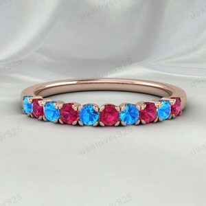 Peut inclure: Bague en or rose ornée de pierres précieuses bleues et rouges alternées. La bague présente un fin anneau et une rangée de pierres rondes. Les pierres sont espacées de manière uniforme, créant un design coloré et attrayant. La bague est posée sur un fond blanc doux.