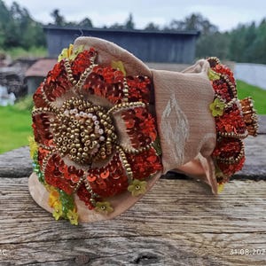 May include: A beige headband featuring a large, decorative floral motif. The flower is crafted from red sequins and gold beads, complemented by small yellow and green embellishments. The headband rests on a weathered wooden surface.