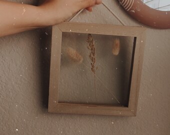 May include: A brown wooden frame with a glass front, holding dried grasses and flowers. The frame is hanging on a light brown wall.