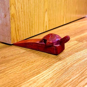 Può includere: Un fermaporta in legno rosso a forma di tartaruga. Il fermaporta è posto su un pavimento in legno, tenendo aperta una porta in legno. La tartaruga ha un guscio e una testa dettagliati.