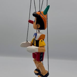 May include: A wooden Pinocchio puppet with a long red nose, wearing a yellow and orange hat, a white collared shirt, and red shorts. The puppet has white gloves and black sandals. It is suspended by black strings.