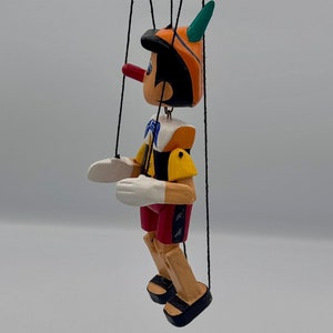 May include: A wooden Pinocchio puppet with a long red nose, orange hat, and yellow shirt. The puppet is suspended by strings from a black wooden frame.