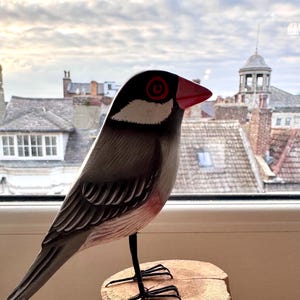 May include: A hand-carved wooden bird figurine perched on a small tree stump. The bird has a black head with a white patch, red beak, and red eye rings. The bird's body is gray with detailed wing markings.