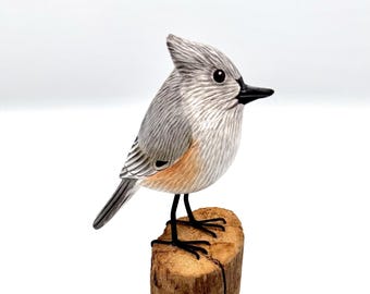 May include: A hand-carved wooden bird figurine, perched on a small wooden block. The bird features gray and white feathers, a black beak, and black legs. The wooden block is a natural brown color.