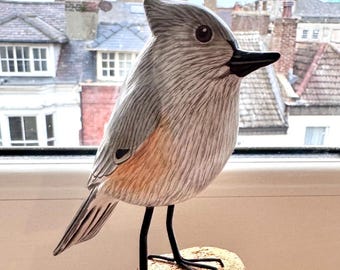 May include: A hand-carved wooden bird sculpture with gray and tan coloring, perched on a small wooden block. The bird has a black beak and legs. The sculpture is displayed near a window with a view of buildings.