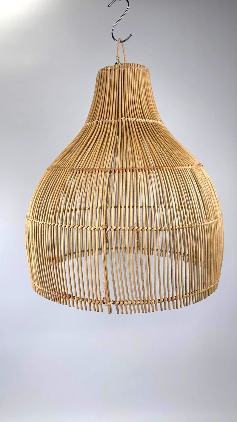 May include: A natural-toned, woven rattan pendant light fixture. The light has a bell-shaped design with a wide base and a narrower top, suspended by a metal hook. The fixture is made of vertical rattan strips.