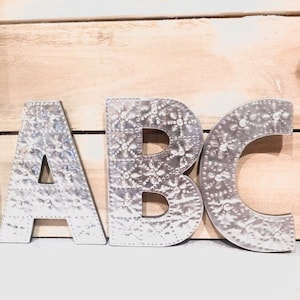 Puede incluir: Letras de metal plateado que deletrean "ABC" con un patrón en relieve decorativo. Las letras están sobre un fondo de madera claro.