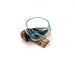 Op de afbeelding: Een heldere glazen kom met een turquoise rand, gevuld met donker, getextureerd materiaal, staat op een stuk drijfhout. Het ontwerp van de kom bevat vloeiende turquoise accenten in het donkere interieur, wat een uniek decoratief stuk creëert.