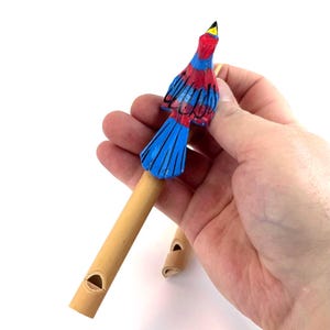 May include: A hand-carved wooden bird whistle with a red, blue, and yellow painted design. The bird is perched atop a light brown bamboo tube with a whistle hole. A second whistle is visible in the background.