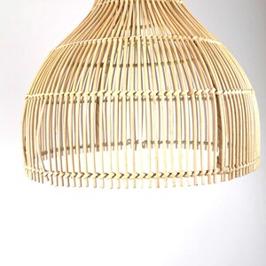 May include: A natural-toned, woven rattan pendant light fixture. The light has a bell-shaped design with a wide base and a narrower top, featuring vertical rattan strips held together by horizontal weaving. The fixture is suspended from a metal hook.
