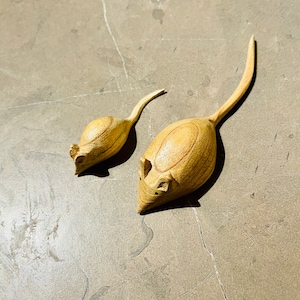 Op de afbeelding: Twee houten muisbeeldjes. De ene is groter dan de andere. Beide zijn gesneden uit lichtgekleurd hout, met lange, gebogen staarten en kleine, gedetailleerde oren. De muizen staan op een grijze, getextureerde ondergrond.