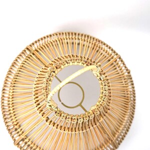 May include: A woven rattan lampshade with a circular design. The lampshade features a natural beige color with a decorative floral pattern around the top. The frame is a light gold color.