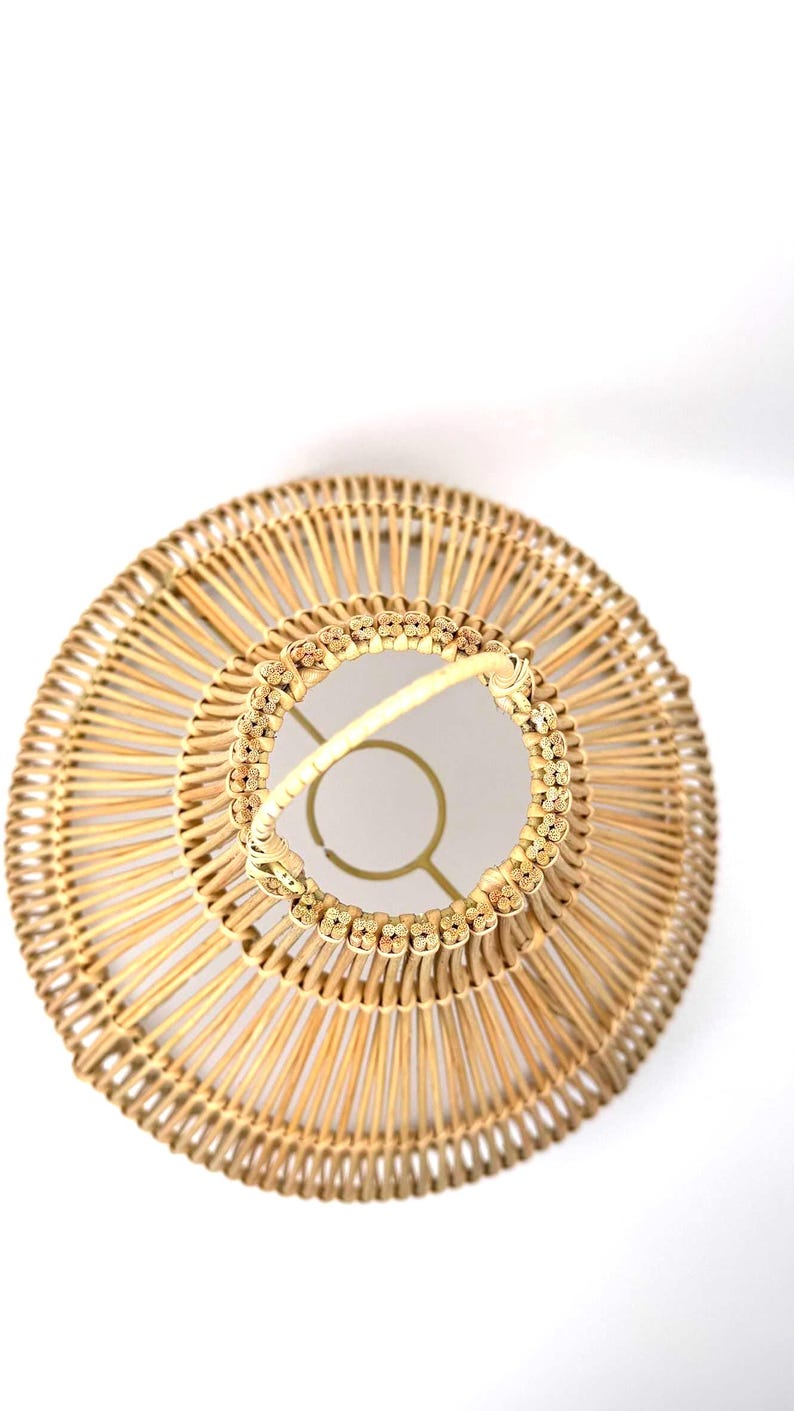 May include: A woven rattan lampshade with a circular design. The lampshade features a natural beige color with a decorative floral pattern around the top. The frame is a light gold color.