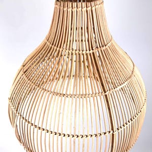 May include: A natural-toned, woven rattan pendant light fixture. The light has a bulbous shape with a flared top and a woven handle. The light is suspended from a black metal hook. The fixture is made of vertical and horizontal rattan strips.