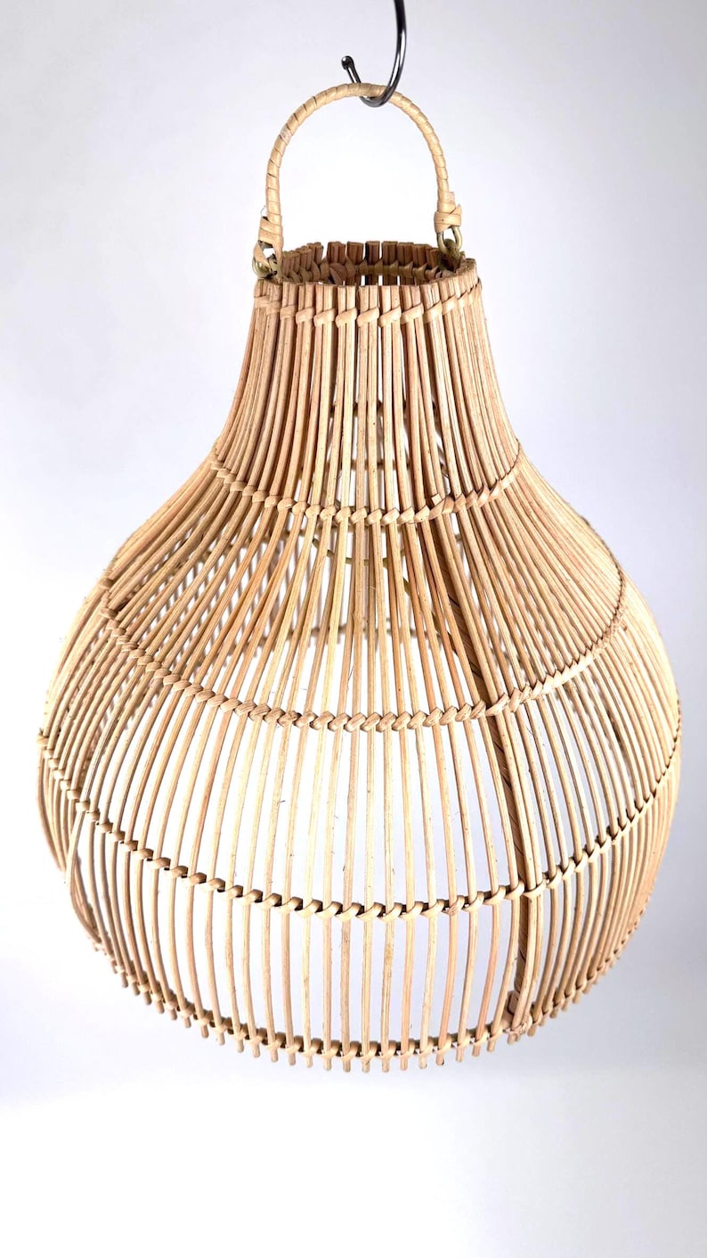 May include: A natural-toned, woven rattan pendant light fixture. The light has a bulbous shape with a flared top and a woven handle. The light is suspended from a black metal hook. The fixture is made of vertical and horizontal rattan strips.