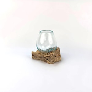 May include: A clear, rounded glass vessel sits atop a piece of weathered driftwood. The glass has a slight blue tint and a wide opening. The wood is a natural brown color with a textured surface. The composition is simple and elegant.