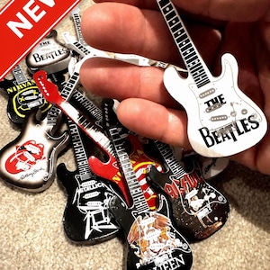 Peut inclure: Collection d'aimants miniatures en forme de guitare, représentant des groupes de rock emblématiques tels que The Beatles, Rolling Stones et Queen. Les guitares sont de différentes couleurs, avec des logos de groupes. Une bannière rouge avec le mot "NEW" est visible.