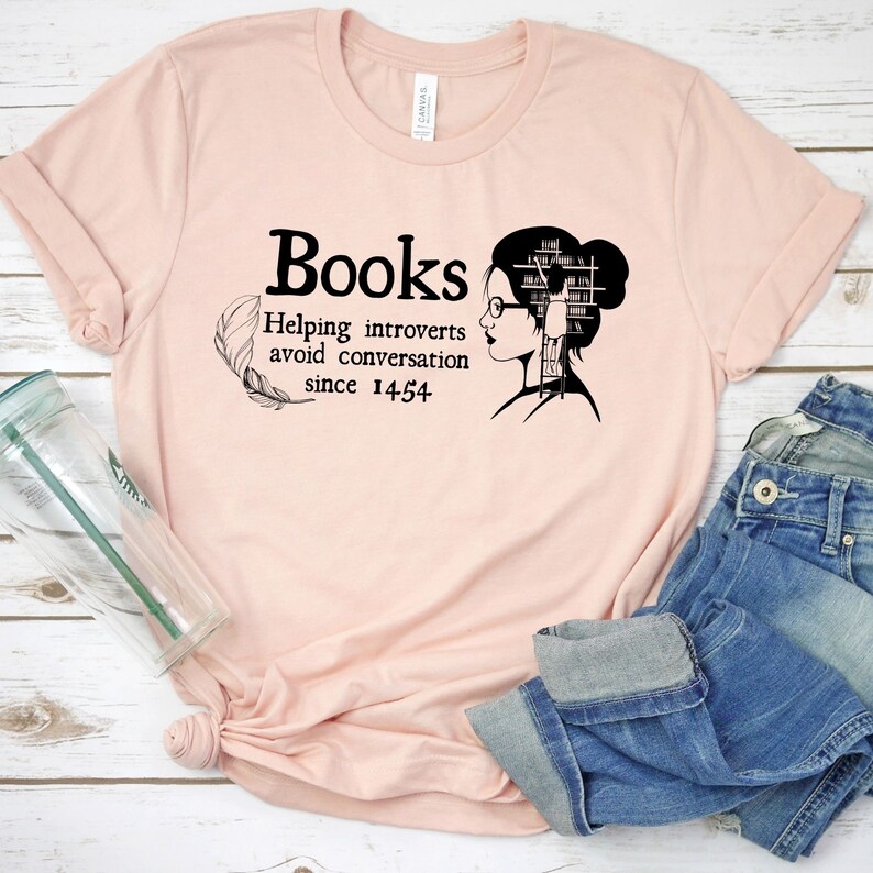May include: A pink t-shirt with a black graphic that says "Books Helping introverts avoid conversation since 1454". The graphic also features a woman with a bookshelf for a head and glasses.