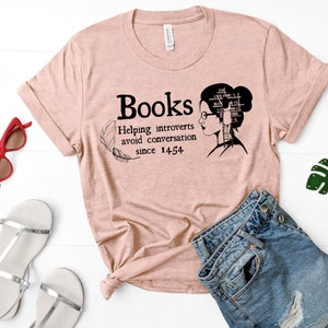 May include: A pink t-shirt with a black graphic that says "Books Helping introverts avoid conversation since 1454". The graphic also features a woman with a stack of books on her head.