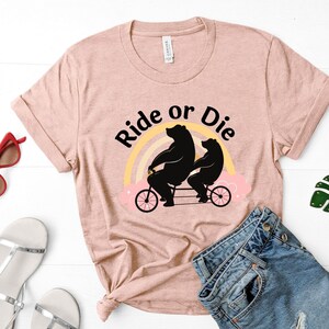 May include: A light pink t-shirt with the words "Ride or Die" above a graphic of two black bears riding a tandem bicycle in front of a rainbow. The shirt is styled with rolled-up sleeves and a knot at the bottom.