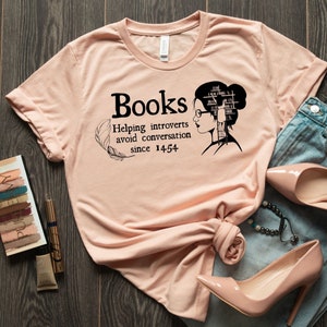 May include: A peach-colored t-shirt with the text "Books Helping introverts avoid conversation since 1454" and a black and white illustration of a woman with books stacked on her head.
