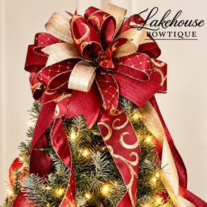 May include: A large decorative Christmas tree topper bow featuring deep red and metallic gold ribbons with intricate patterns, including a lattice design and swirling gold accents, perched atop a lit, flocked evergreen tree. Text reads Lakehouse ROWTIQUE.