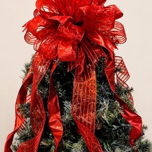 May include: A large, multi-layered red ribbon bow with a sheer, striped texture and glittery accents, positioned atop a flocked evergreen tree. The bow features long, cascading ribbon tails and the text Lakehouse Bowtique in the corner.