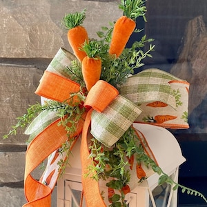 May include: White metal lantern decorated with a large bow featuring orange and green ribbons, faux carrots, and greenery. The ribbons have carrot and gingham patterns, creating a festive Easter theme.