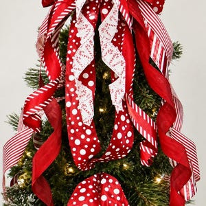 May include: A large decorative Christmas tree topper bow featuring red and white ribbons with various patterns including stripes, polka dots, and candy cane prints. The bow has multiple long, cascading tails and includes the text Lakehouse BOWTIQUE.