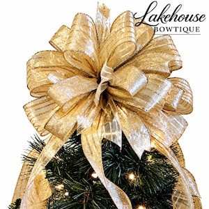 May include: A large, multi-looped gold glitter ribbon bow with sheer striped detailing, positioned atop a dark green pine tree with small warm white lights. The text Lakehouse BOWTIQUE is visible in the upper right corner of the image.