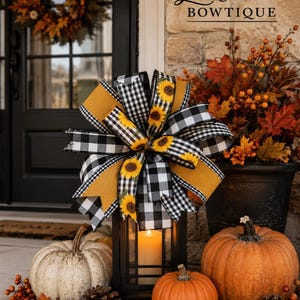 May include: A decorative bow featuring a black and white buffalo check pattern and a sunflower print, accented with solid mustard yellow ribbon. The bow is tied to the top of a black metal lantern containing a single lit pillar candle.