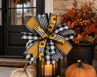 May include: A decorative bow featuring a black and white buffalo check pattern and a sunflower print, accented with solid mustard yellow ribbon. The bow is tied to the top of a black metal lantern containing a single lit pillar candle.
