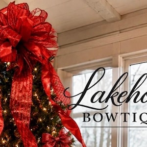 May include: A large, multi-layered red mesh and satin ribbon bow topper sits atop a lit Christmas tree. The text Lakehouse BOWTIQUE is displayed in black script and serif lettering to the right of the tree against a window background.