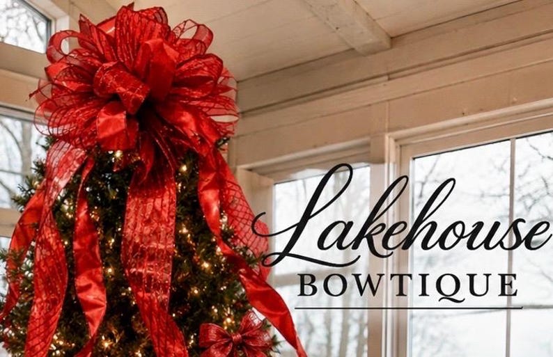 May include: A large, multi-layered red mesh and satin ribbon bow topper sits atop a lit Christmas tree. The text Lakehouse BOWTIQUE is displayed in black script and serif lettering to the right of the tree against a window background.