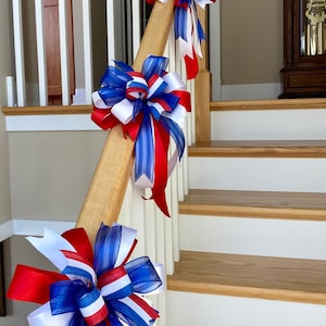 May include: Red, white and blue ribbon bows are tied to a wooden handrail on a staircase. The bows are made of sheer fabric and have a striped pattern.