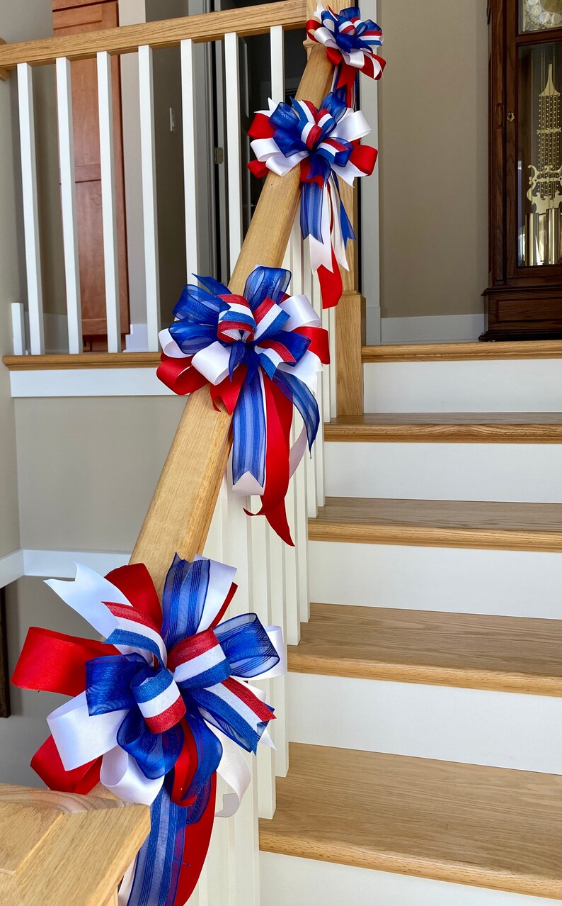 May include: Red, white and blue ribbon bows are tied to a wooden handrail on a staircase. The bows are made of sheer fabric and have a striped pattern.