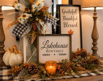 May include: A decorative display featuring a white sign with the text Lakehouse BOWTIQUE, a taller sign with the text Grateful Thankful Blessed, a white pumpkin, a candle, pinecones, greenery, and a large bow with a sunflower and plaid pattern.